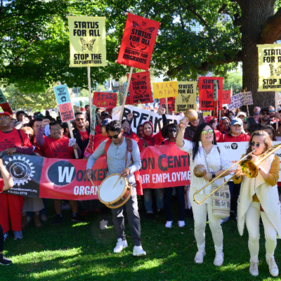 WAC members at the Draw the Line action hold their red Workers' Action Centre banner and red and yellow placards that say "Status for All"