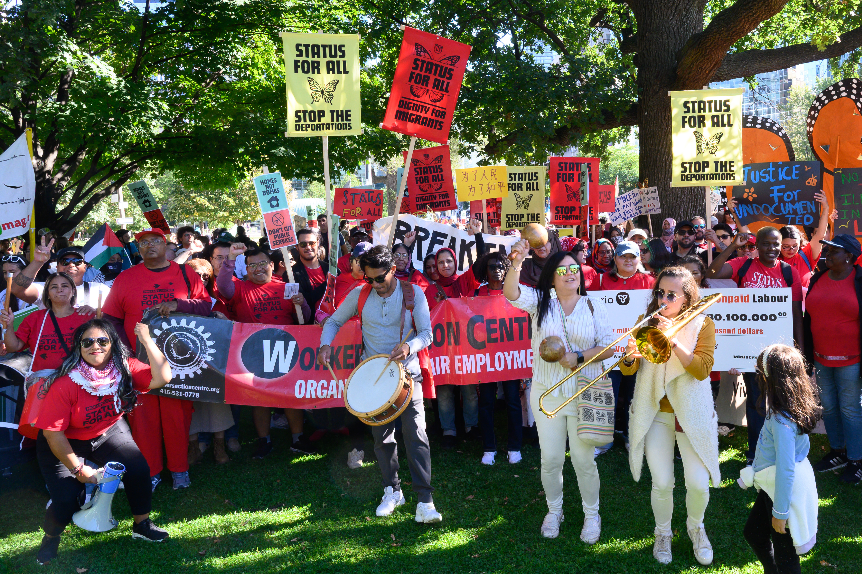WAC members at the Draw the Line action hold their red Workers' Action Centre banner and red and yellow placards that say "Status for All"